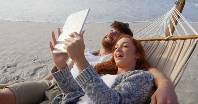 Couple enjoying leisure time on beach hammock video chatting on tablet near ocean. Ideal for vacation themes, travel promotions, technology usage, couple bonding, and leisure activities.