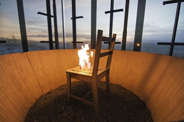 Wooden chair engulfed in flames inside a circular structure with glass walls overlooking an evening horizon. Flames create a dramatic and evocative scene, making this suitable for themes of destruction, artsy designs, rebellion, or contemporary issues. Ideal for editorial use, artistic projects, and thought-provoking advertisements.