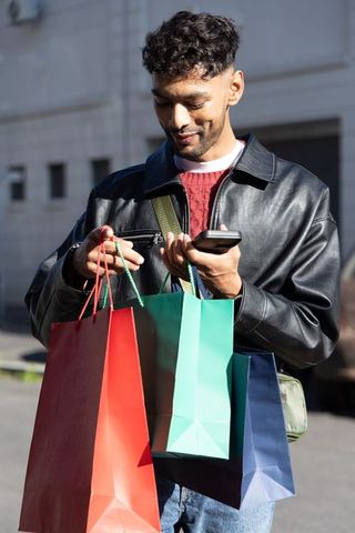 Urban asian man holding colorful shopping bags while checking phone in leather jacket