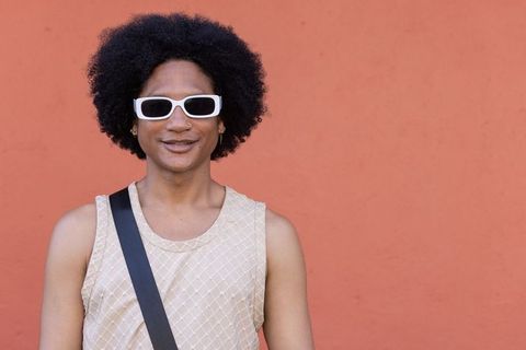 Urban stylish young man wearing white sunglasses standing against terracotta wall with crossbody bag