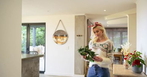 Woman Decorating with Lit Evergreen Wreath in Modern Sunlit Home Wearing Festive Sweater
