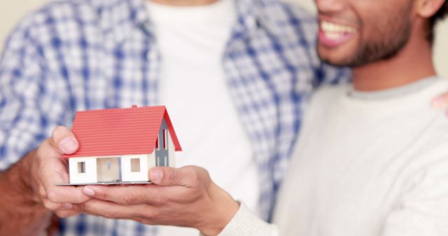 Happy friends smiling while holding a miniature house model symbolizing new homeownership. Ideal for content about real estate, property purchase, first-time home buyers, housing market, investment, home celebrations, and community support.