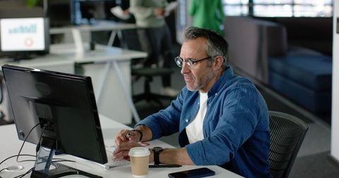 Mature professional focusing on computer at open-plan office desk with coffee, smartphone