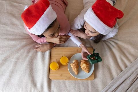 Diverse women wearing santa hats sharing cozy holiday breakfast in bed with croissants