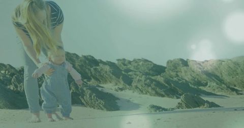 Young woman steadying baby learning to walk on sunlit rocky beach in denim overalls