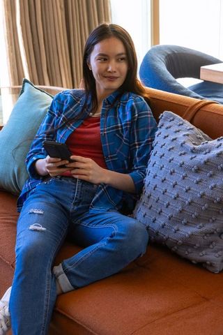Relaxed asian woman holding smartphone on comfortable orange sofa