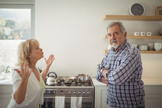 Senior couple having a heated argument in a modern kitchen. The woman is gesturing with her hands while the man stands with his arms crossed, looking away. This image can be used in articles or advertisements related to family dynamics, relationship counseling, elderly care, or domestic life.