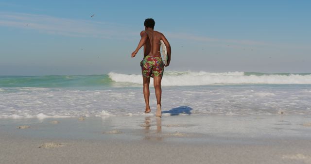 Young man running towards the ocean carrying a surfboard on a sunny beach. Ideal for travel brochures, outdoor lifestyle blogs, summer vacation promotions, or advertisements related to water sports and beach activities.