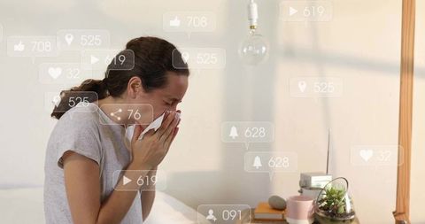 Mid-adult woman sneezing in bed using tissue with social media notification overlays