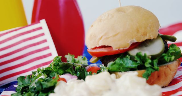 A close-up of a classic American hamburger with fresh vegetables and condiments, with copy space. The setting suggests a festive atmosphere, a barbecue or a Fourth of July celebration.