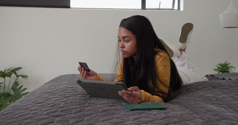 African american woman lying on bed shopping online with tablet and credit card, modern lifestyle