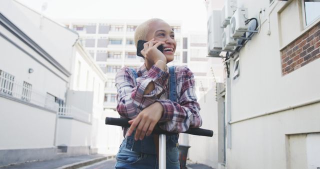 Woman with short hair is riding a scooter while talking on the phone in an urban alley. She is dressed casually in a plaid shirt and is smiling cheerfully. This visual can be perfect for representing communication style, carefree lifestyle, city adventures, or urban daily life.