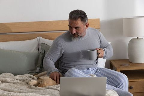 Mature man sipping coffee and petting dog while working on laptop in cozy bedroom