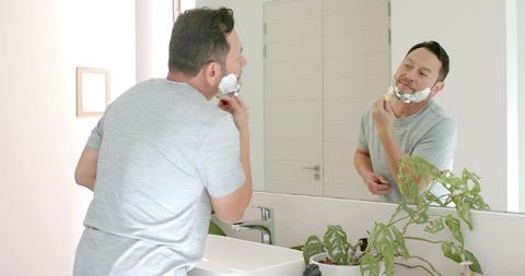 Mature man shaving with razor in bathroom reflection