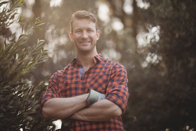 Young man standing confidently by an olive tree at a farm, wearing a plaid shirt and smiling with arms crossed. Ideal for use in agricultural promotions, rural lifestyle blogs, or outdoor activity advertisements.