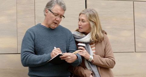Senior couple planning trip together outdoors: man writing notes while woman holding coffee