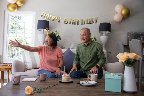 Asian senior couple celebrating birthday in living room, woman reaching for cake