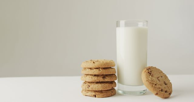 Glass of milk with stack of chocolate chip cookies placed on white table. Perfect for usage in food blogs, recipe websites, or advertisement for dairy or baking products. This image evokes a sense of comfort and nostalgia, making it ideal for promoting snack-time items or homemade bakery goods.