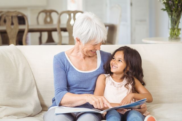 Grandmother and granddaughter sitting on sofa, looking at photo album, smiling and bonding. Ideal for family-oriented content, advertisements promoting family values, and articles about intergenerational relationships.