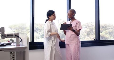 Black Nurse Reviewing Tablet With Asian Patient in Gown During Clinical Consultation