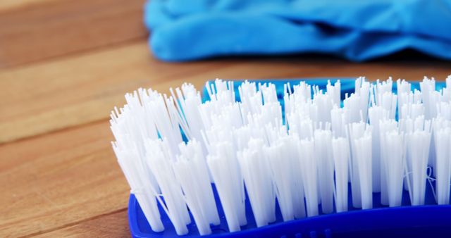Horizontal color photo showing a close-up cleaning brush with white bristles on a wooden surface with a blue glove in the background. This image is useful for illustrating cleaning products, hygiene themes, and household maintenance activities in articles, blogs, and advertisements.