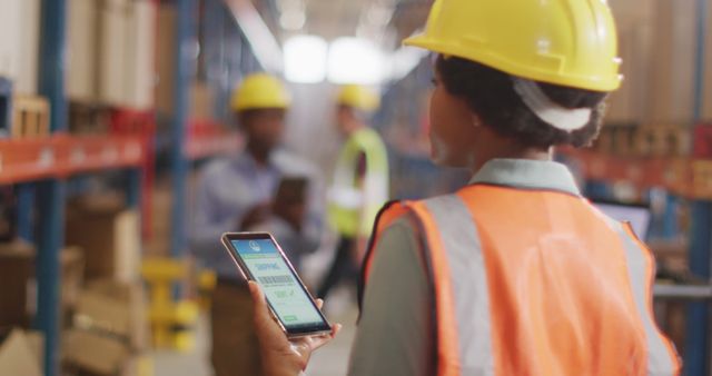 African american female worker with helmet using smartphone in warehouse. global business, shipping and delivery.