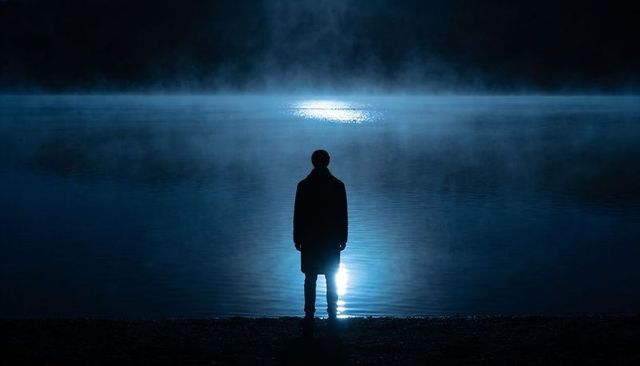 Solitary silhouette standing at misty night lake with vertical moonlit reflection