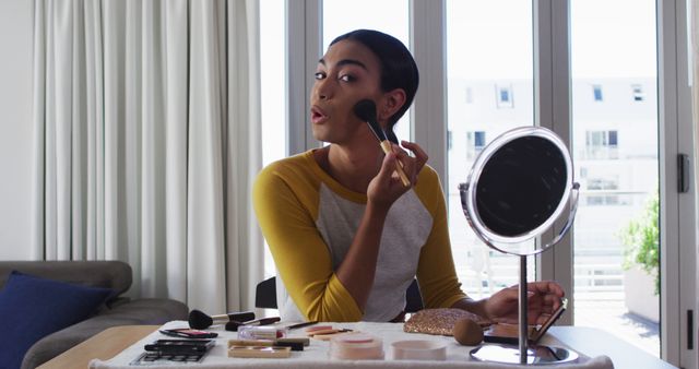 Young woman applying makeup with brush at home, seated at table with various beauty products. Suitable for content on beauty, skincare routines, lifestyle, and home self-care.