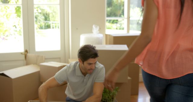 Scene shows a couple engaged in the moving process inside their new home. The man is kneeling and packing a plant into a cardboard box, while the woman walks across the room. There are several boxes scattered around, and the room is well-lit with sunlight coming through large windows. Useful for concepts related to moving, relocation, new beginnings, and home organization.