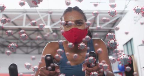 Woman wearing red face mask cycling on stationary bike with floating virus graphics in gym