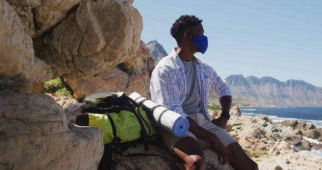 African american man wearing face mask hiking in mountain countryside sitting on a rock. fitness training and healthy outdoor lifestyle during coronavirus covid 19 pandemic.
