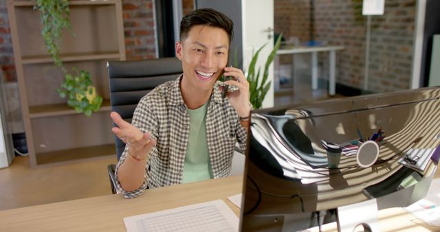 Young professional man is talking on the phone while sitting at an office desk. He is using various gadgets and is in a modern office environment with plants and brick walls. This can be used for themes relating to business, office work, productivity, technology, and modern workspaces.