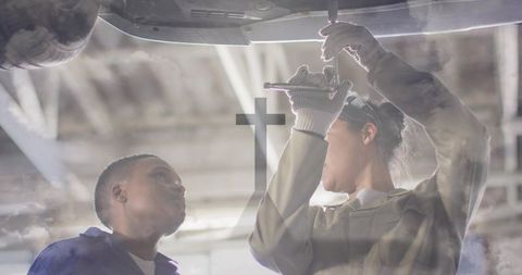 Female Technician Mentoring Junior Mechanic While Repairing Car Under Hoist in Workshop