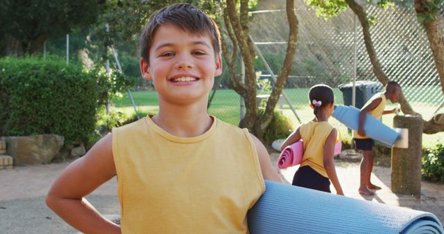 Boy carrying blue yoga mat while smiling at outdoor fitness session during sunny day. Children in background engaging in physical activity with yoga mats. Suitable for themes related to childhood health, outdoor activities, fitness programs for children, and summer camps.