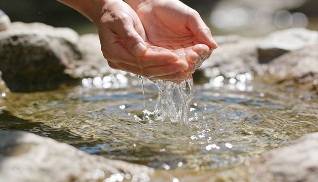 Cupped hands pouring clear spring water over sunlit rock pool closeup