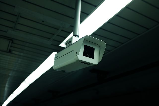 Security camera mounted on a ceiling in what appears to be an industrial environment. The camera is white and aligned under bright fluorescent lights. This image can be used for themes related to security, surveillance technology, industrial safety, and monitoring systems. Ideal for use in articles about security measures, facility safety, and smart technology solutions.