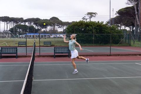 Female tennis player serving on outdoor court in athletic polo and skirt, midair serve