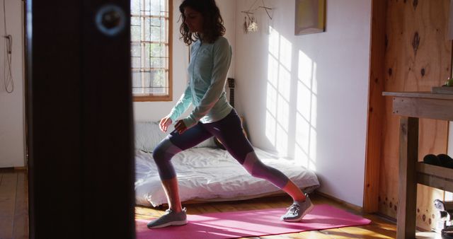 Woman performing a fitness routine in a bright and sunlit room, utilizing a yoga mat. Ideal for content related to home workouts, fitness routines, wellness tips, healthy lifestyles, and the benefits of indoor exercise spaces.