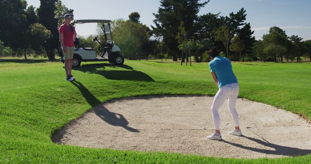 Two caucasian women playing golf wearing face masks one taking shot from bunker. golf cart and clubs in background. golf sports hobby healthy lifestyle hygiene during covid coronavirus pandemic.