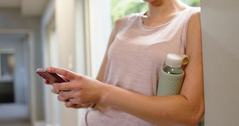 Leaning woman checking phone in hallway holding mint water bottle casual tank top