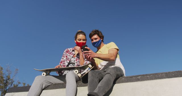 Caucasian woman and man friend wearing face mask, sitting and using smartphone. hanging out at skate park in summer during coronavirus covid 19 pandemic.