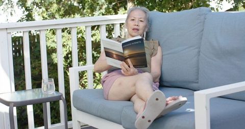 Senior woman reclining on blue outdoor sofa reading paperback, relaxing on sunlit deck