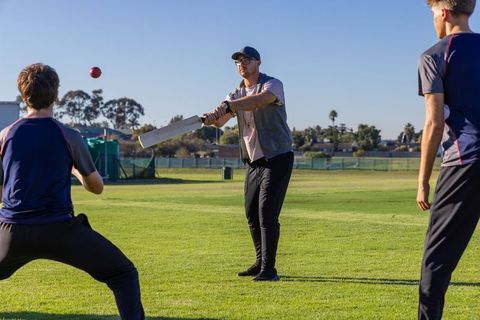 Coach batting with teammates on sunlit grass cricket field during training session