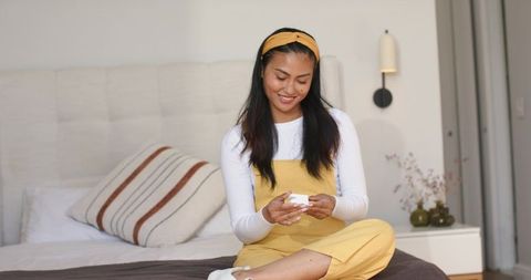 Smiling asian woman sitting cross-legged on bed holding small gift box in yellow outfit