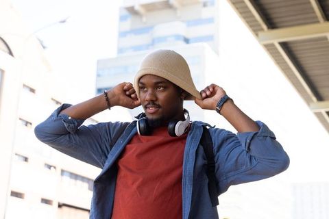 African american man adjusting beanie while wearing headphones on city sidewalk