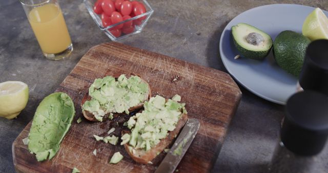 Image shows avocado toast preparation in a modern kitchen. Fresh ingredients including avocados, lemon, cherry tomatoes, and a glass of orange juice are visible. Ideal for blogs on healthy eating, food magazines, cooking tutorials, or healthy lifestyle promotions.