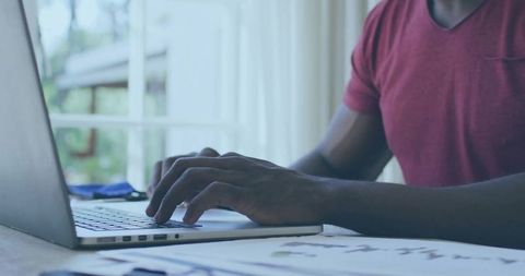 Man typing on laptop at home desk with documents and glasses in natural daylight home office