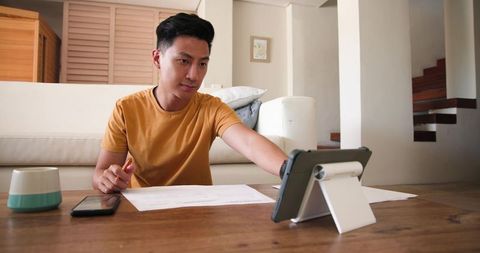 Man Studying Documents at Home While Reaching for Tablet on Table