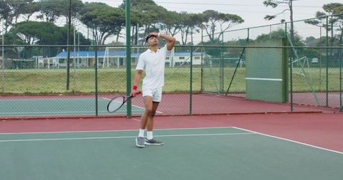 Asian tennis player shading eyes while holding racket on outdoor hard court