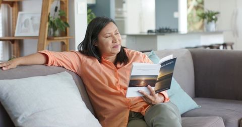 Mature asian woman reading paperback on cozy gray sofa in sunlit living room